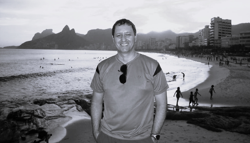 Fotografia em preto e branco de um homem. Ele está em pé, com as mãos nos bolsos e sorri para a câmera. Atrás dele há uma paisagem com uma praia, alguns prédios e morros.
