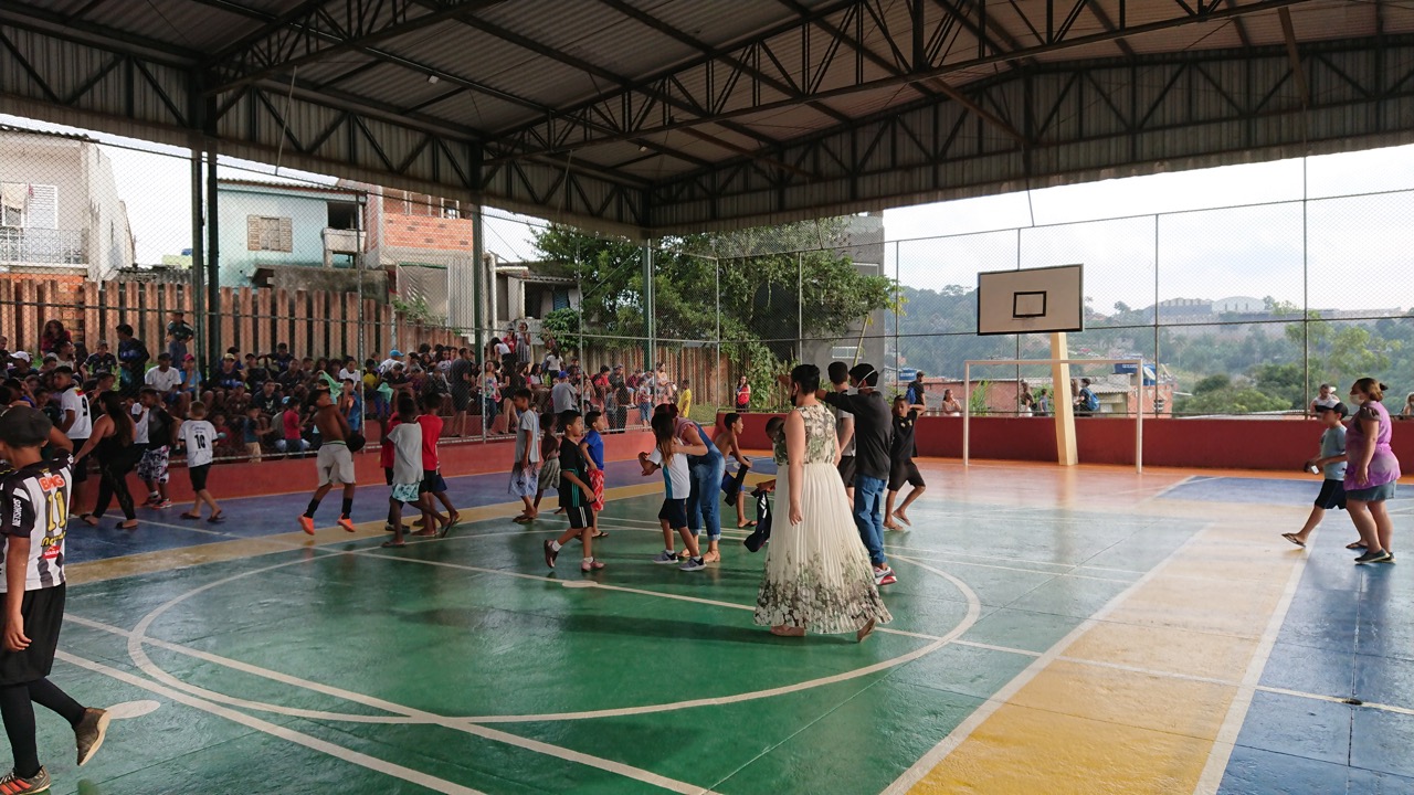Fotografia de vários adultos e crianças reunidos em uma quadra poliesportiva escolar pintada de verde, amarelo, azul e laranja.