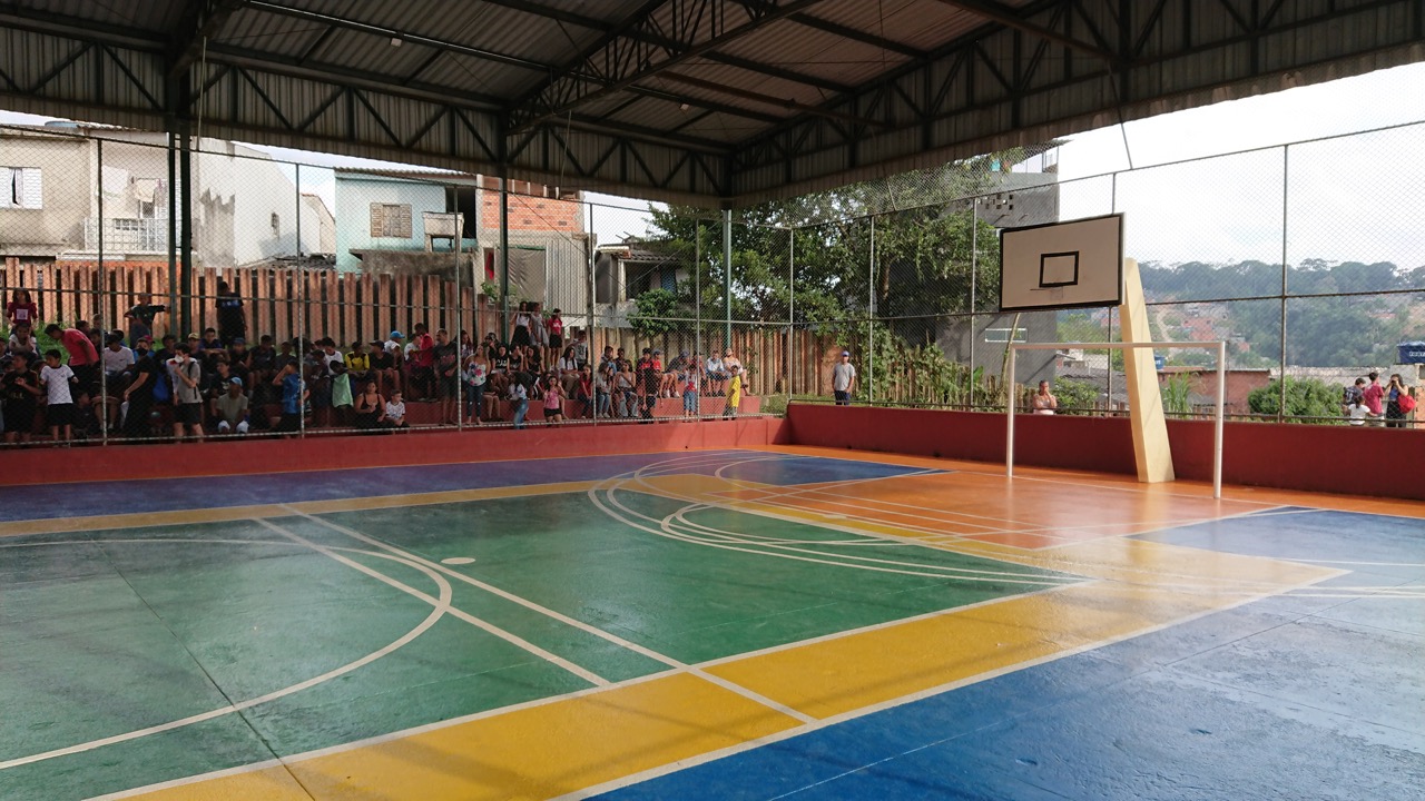 Fotografia de quadra poliesportiva escolar vazia e pintada de verde, amarelo, azul e laranja. Ao fundo, várias pessoas estão reunidas em uma arquibancada.