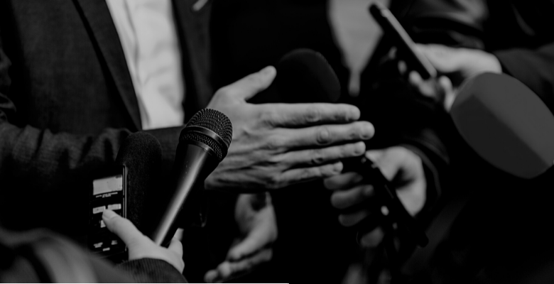 Fotografia em preto e branco que foca em diversos microfones e gravadores diante de uma pessoa que está sendo entrevistada.